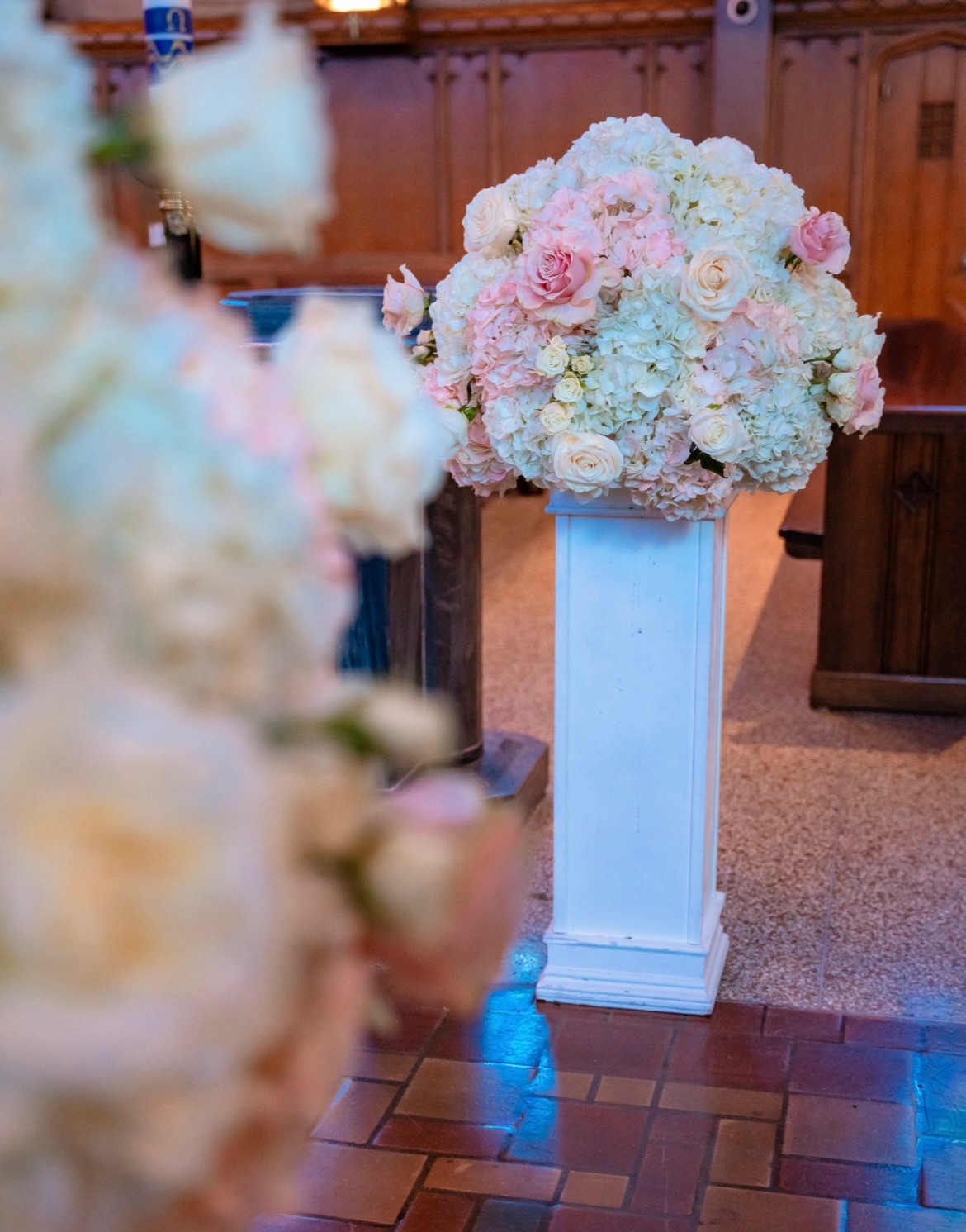 Close view of a blush rose and white hydrangea dome on a white column beside the church pews