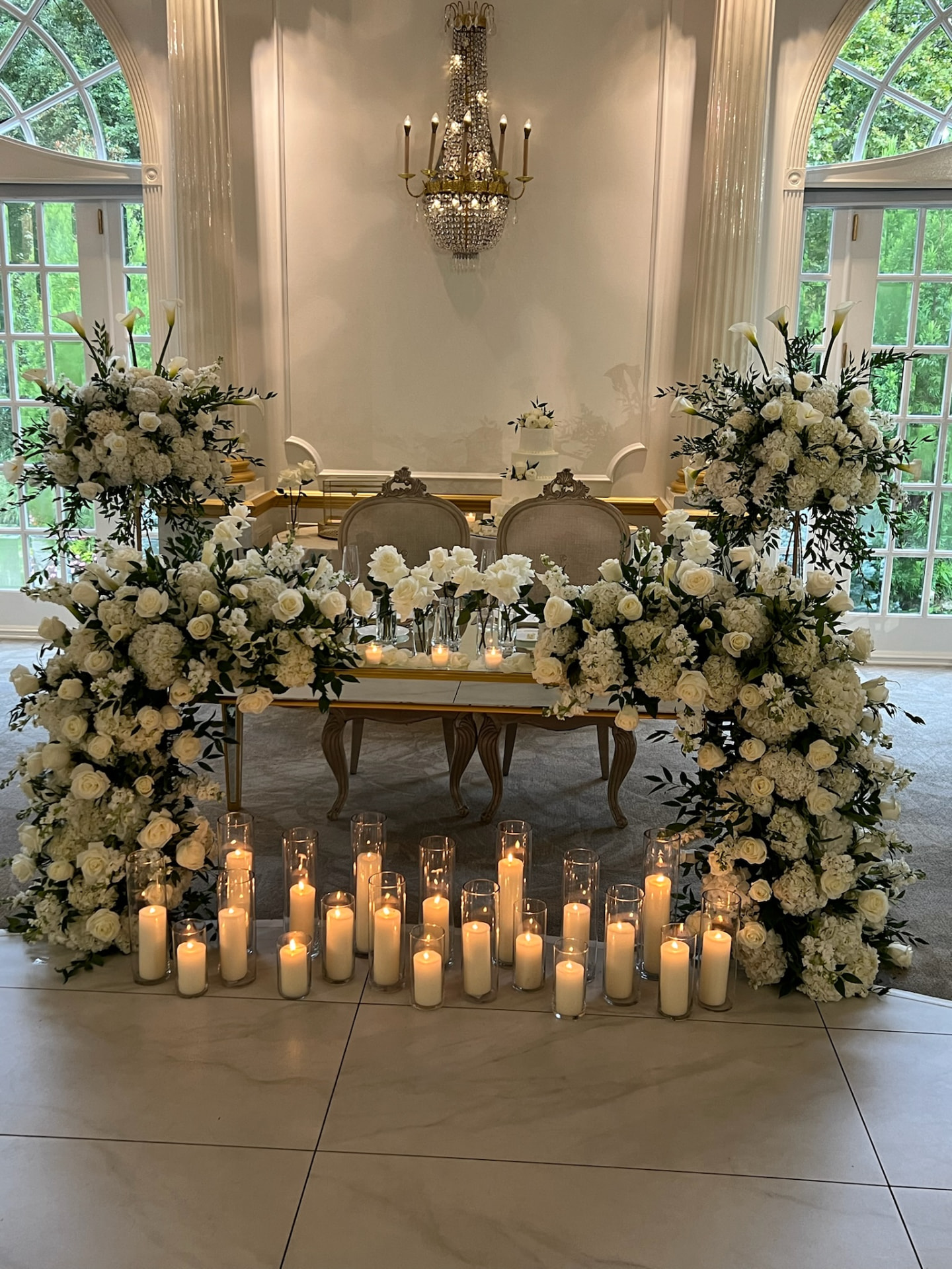 Sweetheart table framed by two white rose and calla lily towers with rows of pillar candles on a marble floor