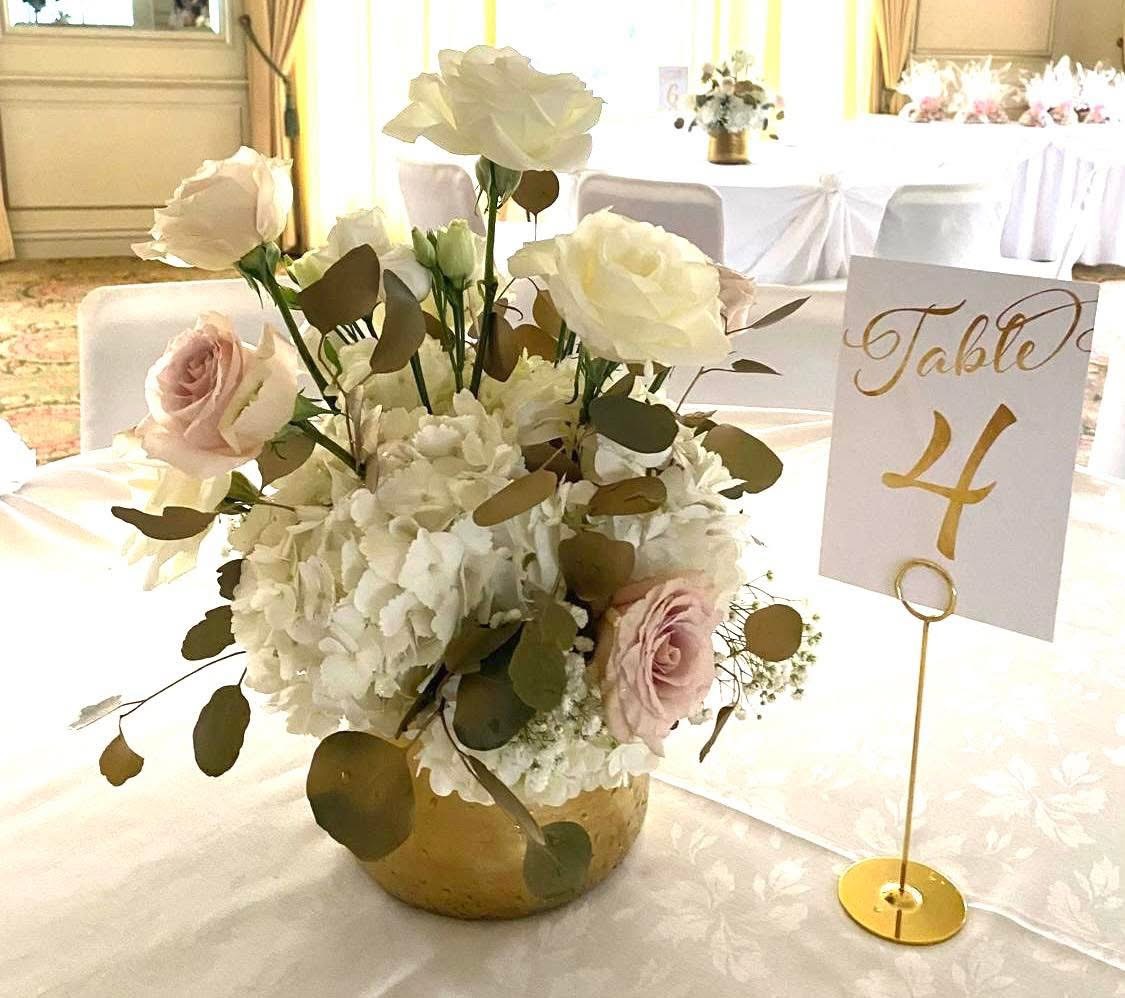 Low centerpiece of white and blush roses with hydrangea and eucalyptus in a gold bowl beside a gold table number