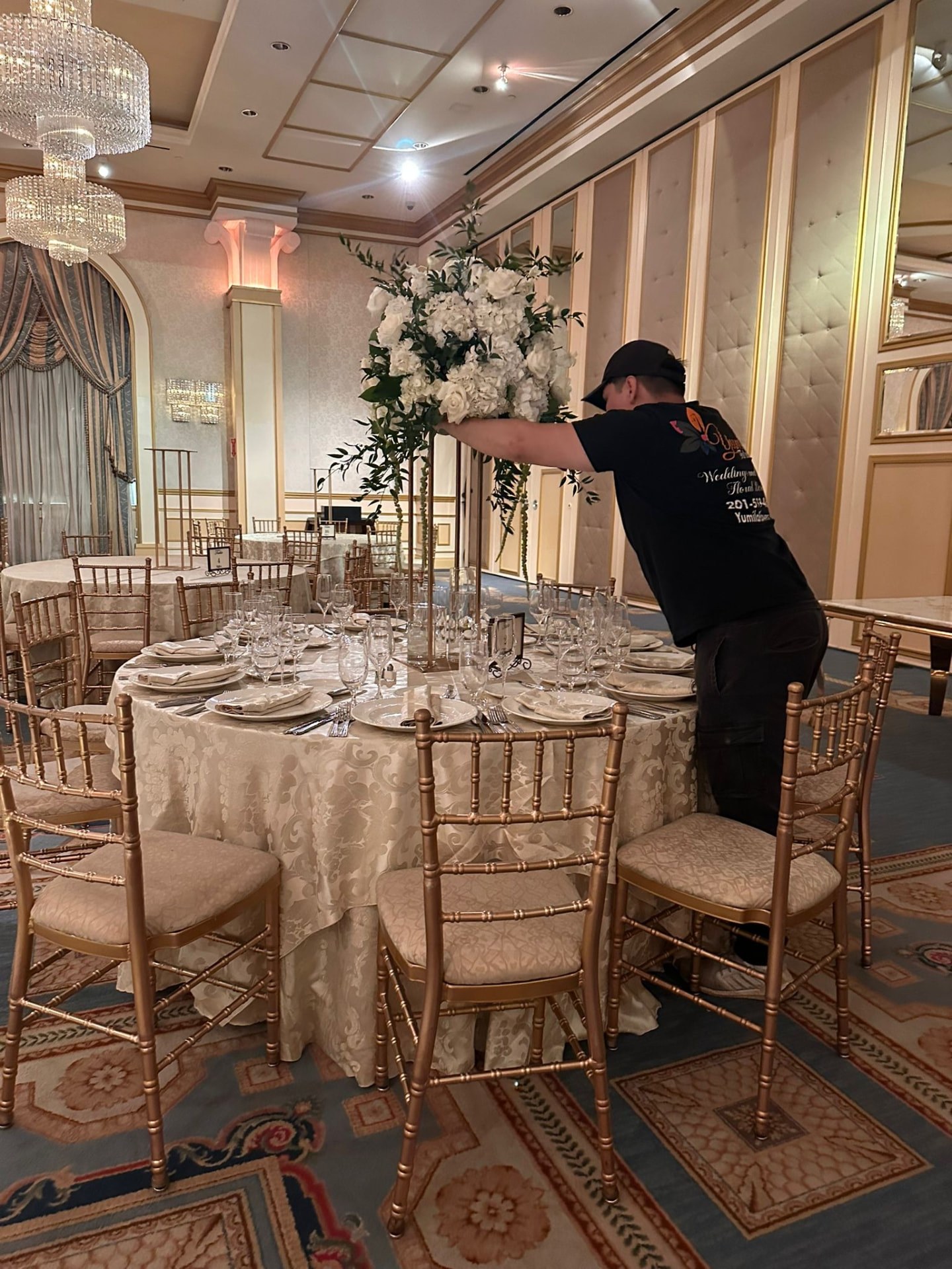 A Yumila’s designer setting an elevated white hydrangea and greenery centerpiece on a gold-set reception table