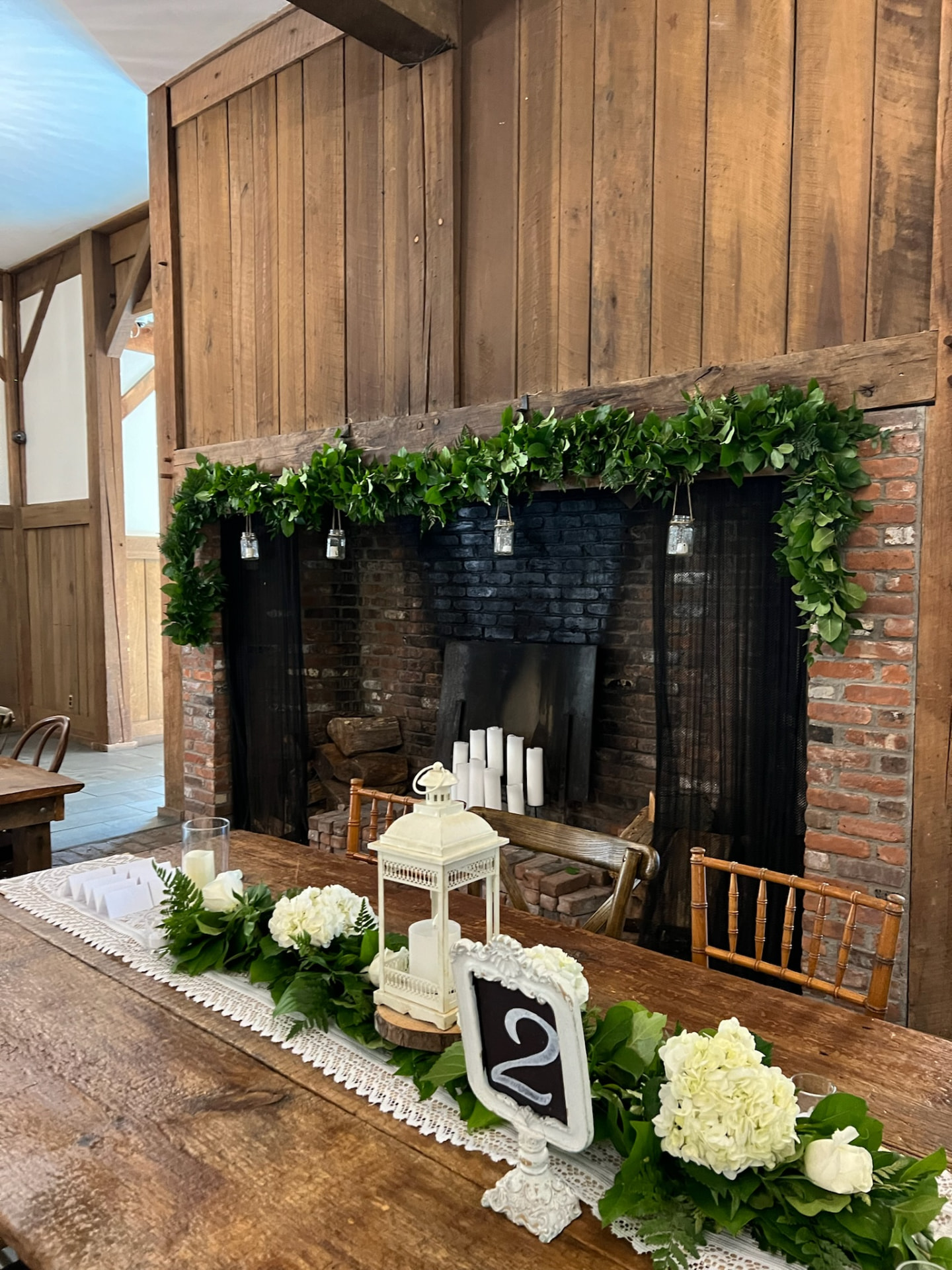 Rustic barn fireplace mantel wrapped in fresh greenery garland above a lace-runner table with hydrangea