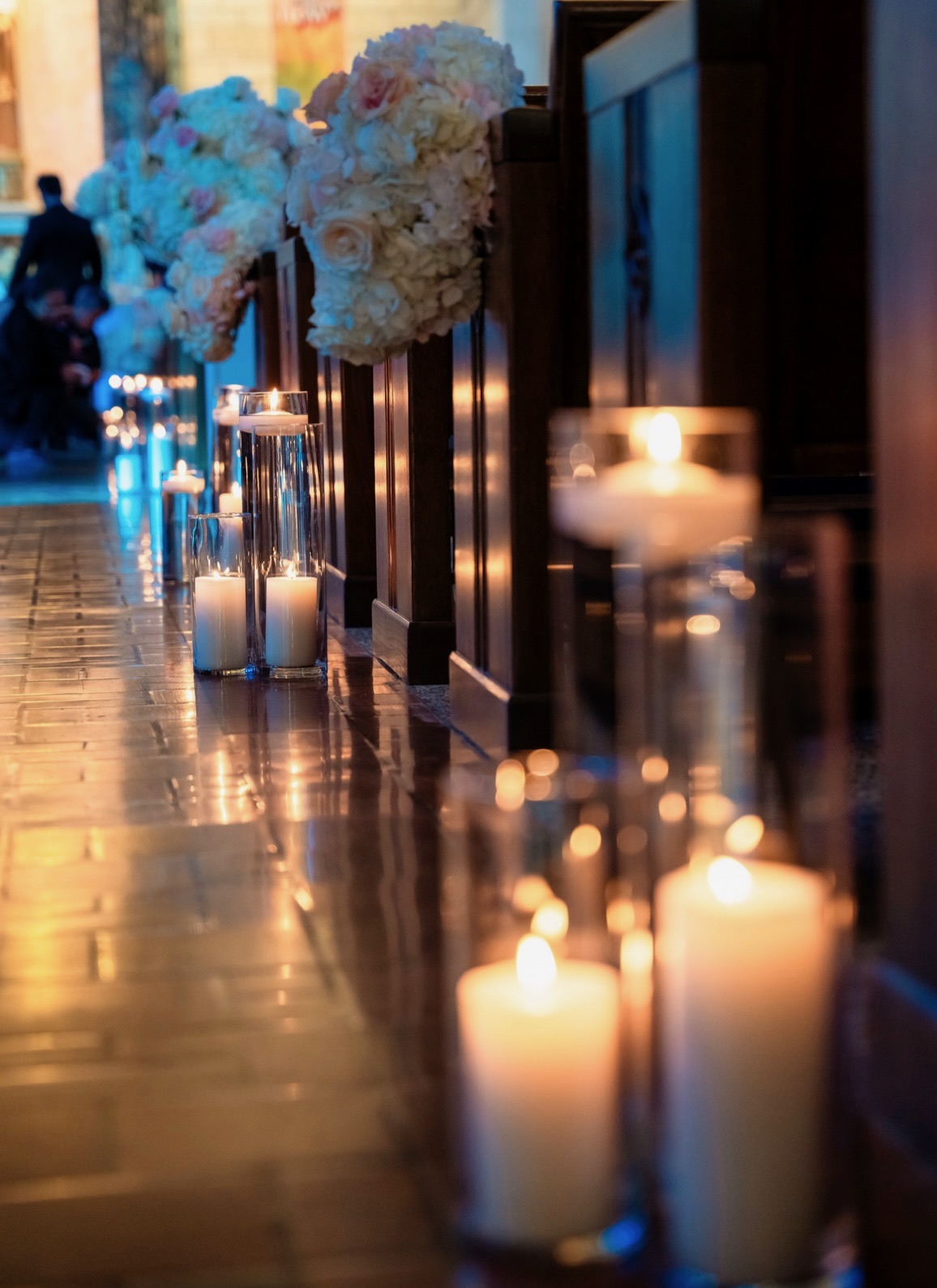 Detail of candle-lined church pews with white rose and hydrangea aisle arrangements above