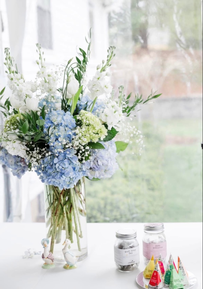 Tall glass vase of blue hydrangea, white stock and bells of Ireland on a tea table by a window
