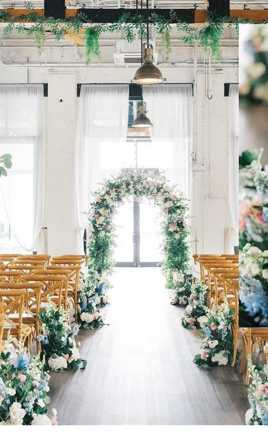Bright brick hall ceremony with a greenery and blush rose arch between rows of wooden cross-back chairs