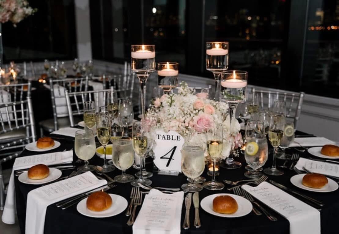 Round table in black linen with a blush and white centerpiece, floating candles, and a table four card