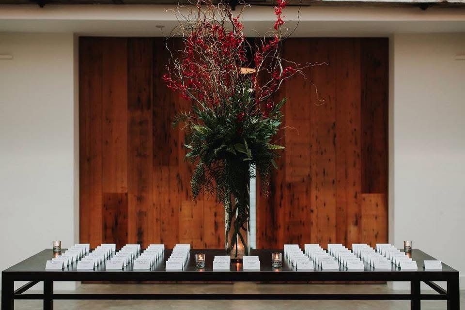 Escort card table with a tall arrangement of red branches and ferns against a wood wall