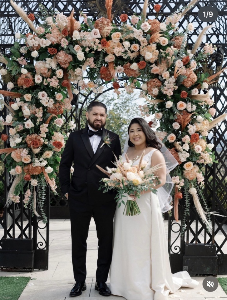 Couple before a terracotta, rust and pampas floral arch on black iron gates