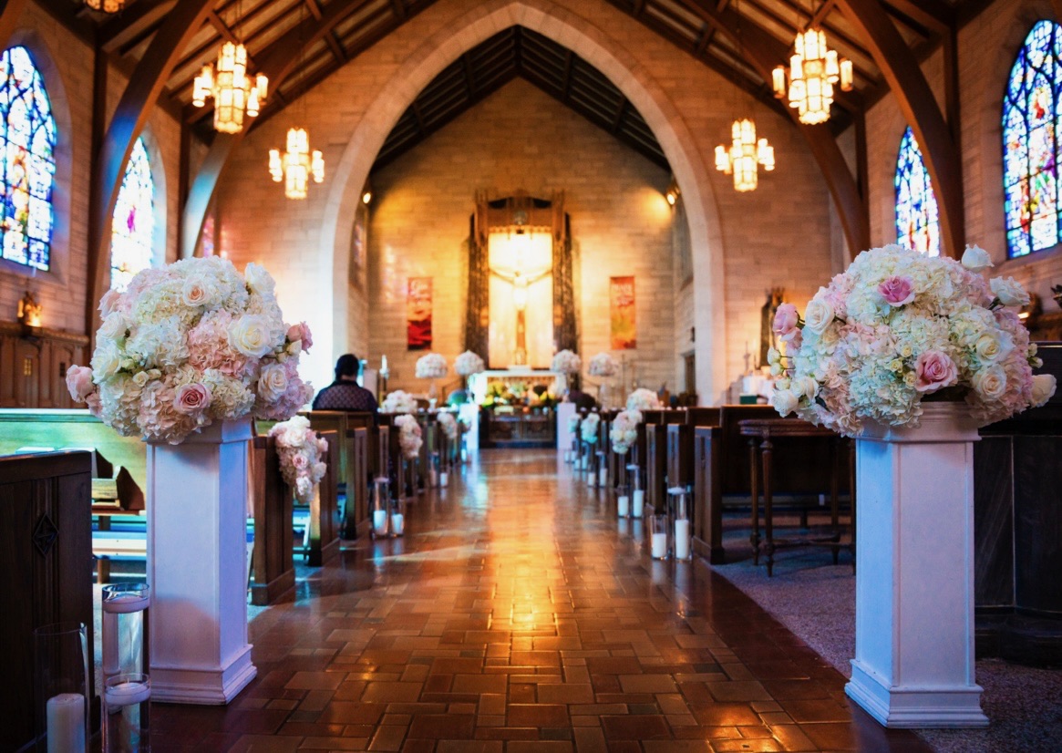 Church aisle framed by white pedestals of blush roses and hydrangea leading to a candlelit altar