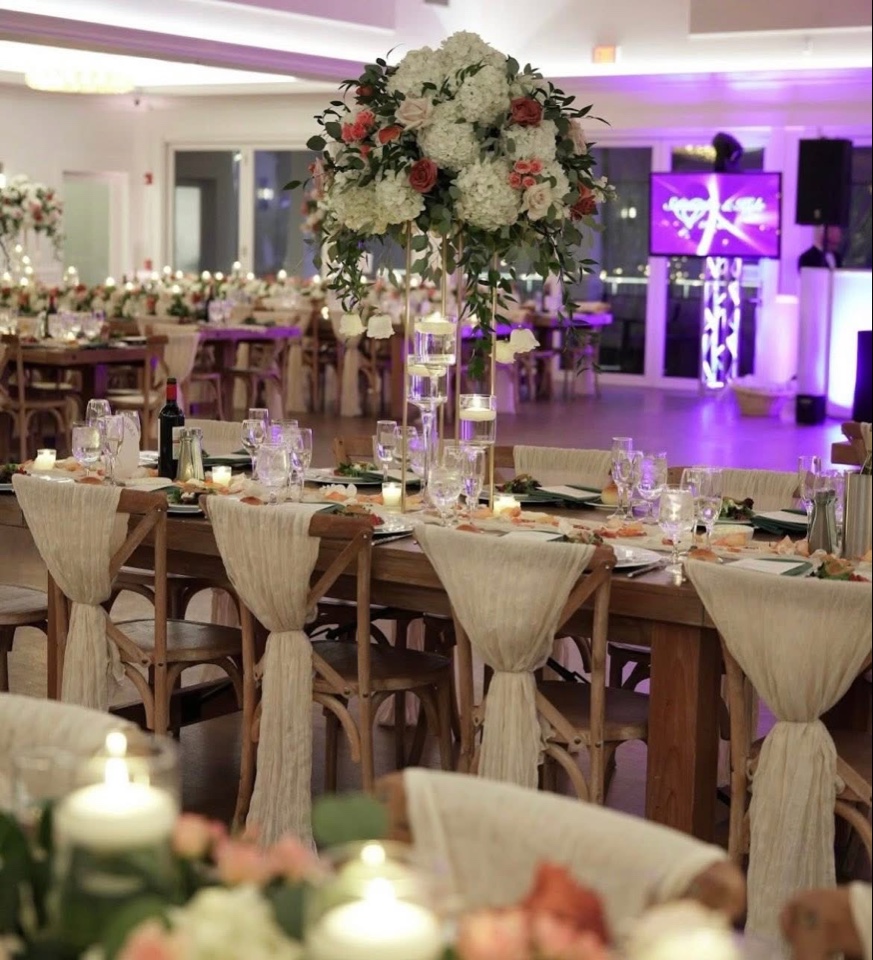 Farm table reception with a tall white hydrangea and coral rose centerpiece and ivory chair sashes