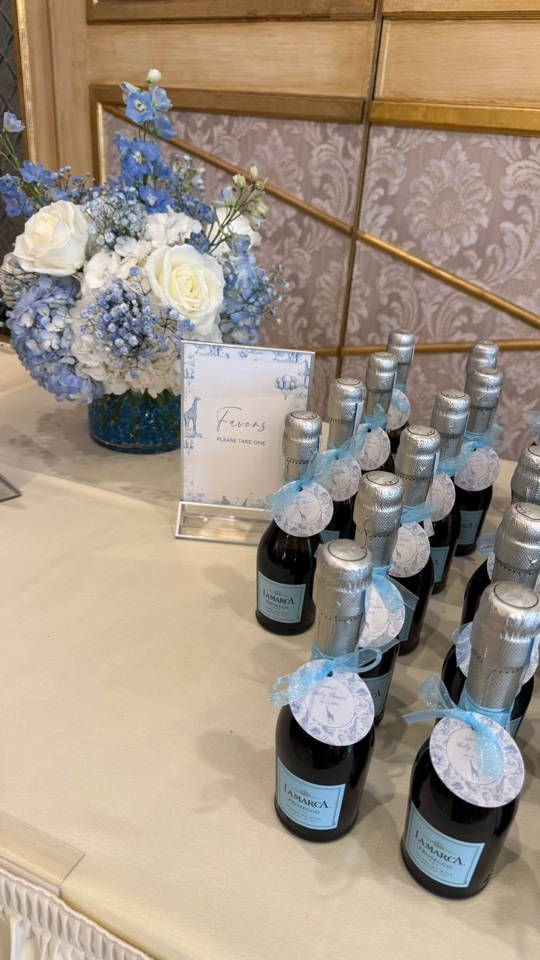 Favors table with blue hydrangea and white rose bouquet beside mini champagne bottles tied with blue toile tags