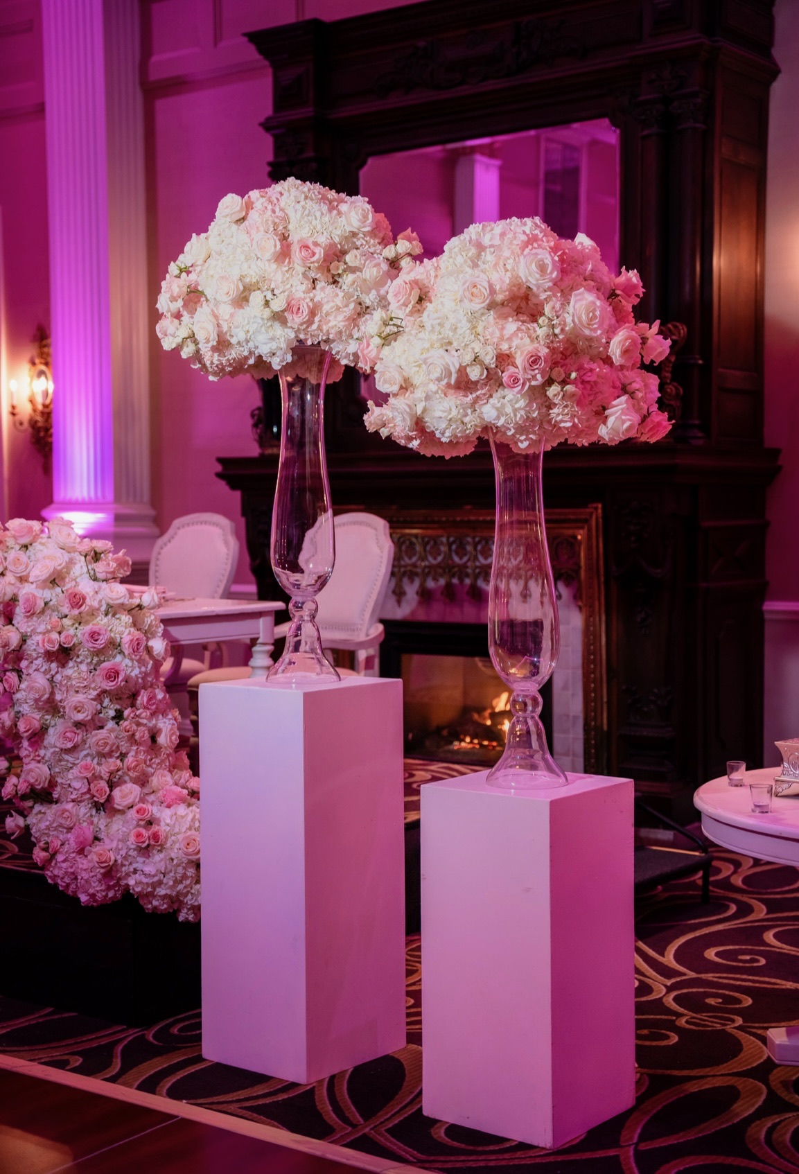 Pair of pedestal vases topped with pink and ivory rose domes beside a fireplace in magenta light