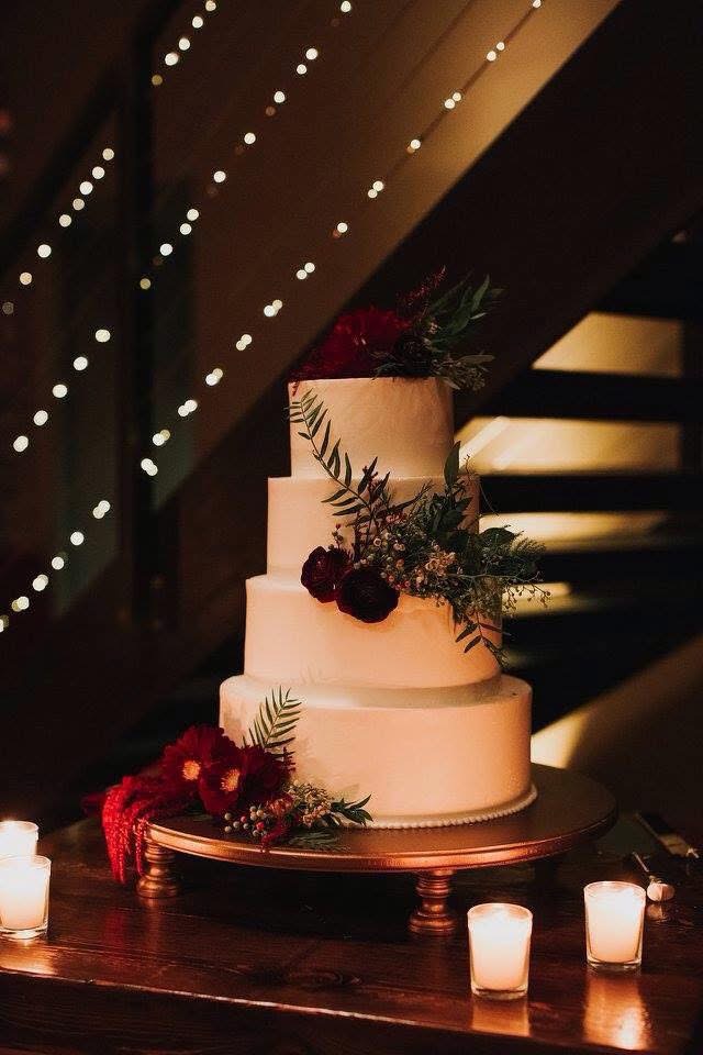 Four-tier wedding cake decorated with burgundy blooms and greenery under string lights