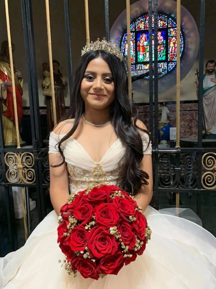 Quinceañera in an ivory gown and tiara holding her red rose bouquet in front of the church stained glass