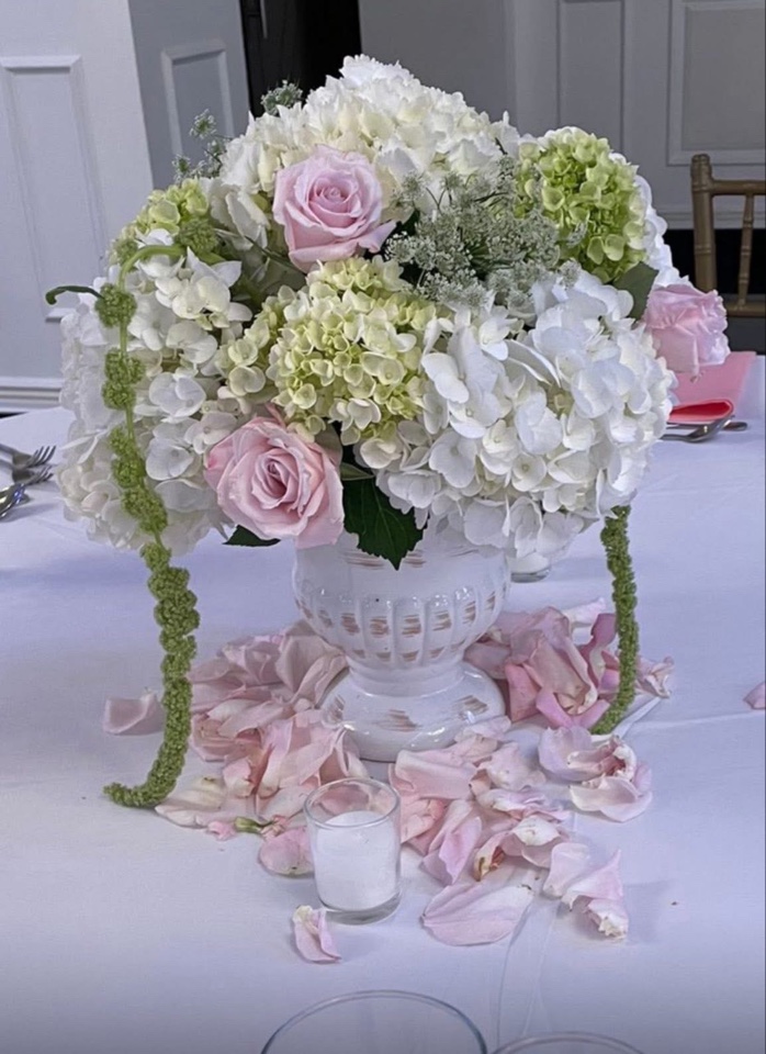 White urn centerpiece of hydrangea and blush roses with trailing greenery and scattered rose petals