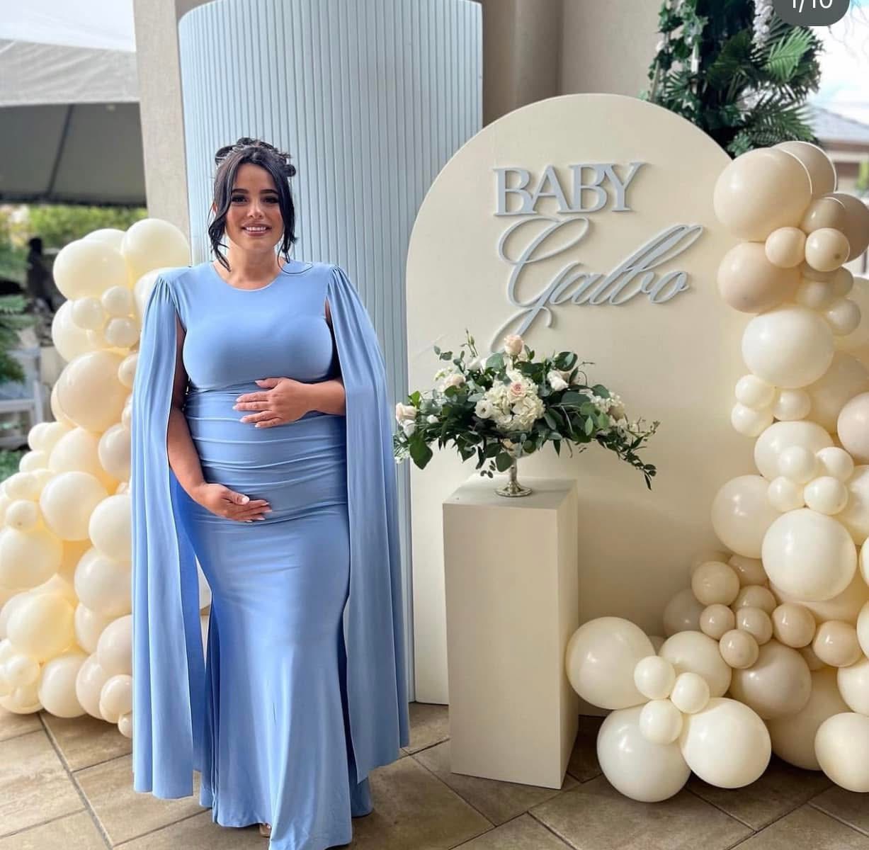 Mom-to-be in a powder blue cape gown beside a Baby Galbo arch with cream balloons and a pedestal of white florals