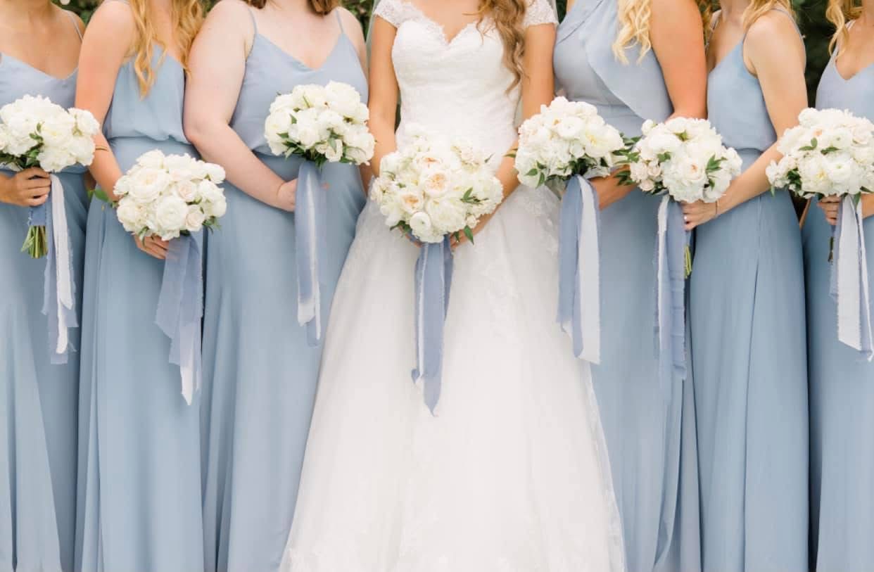 Bride and bridesmaids in dusty blue gowns holding white rose and peony bouquets with trailing blue ribbon