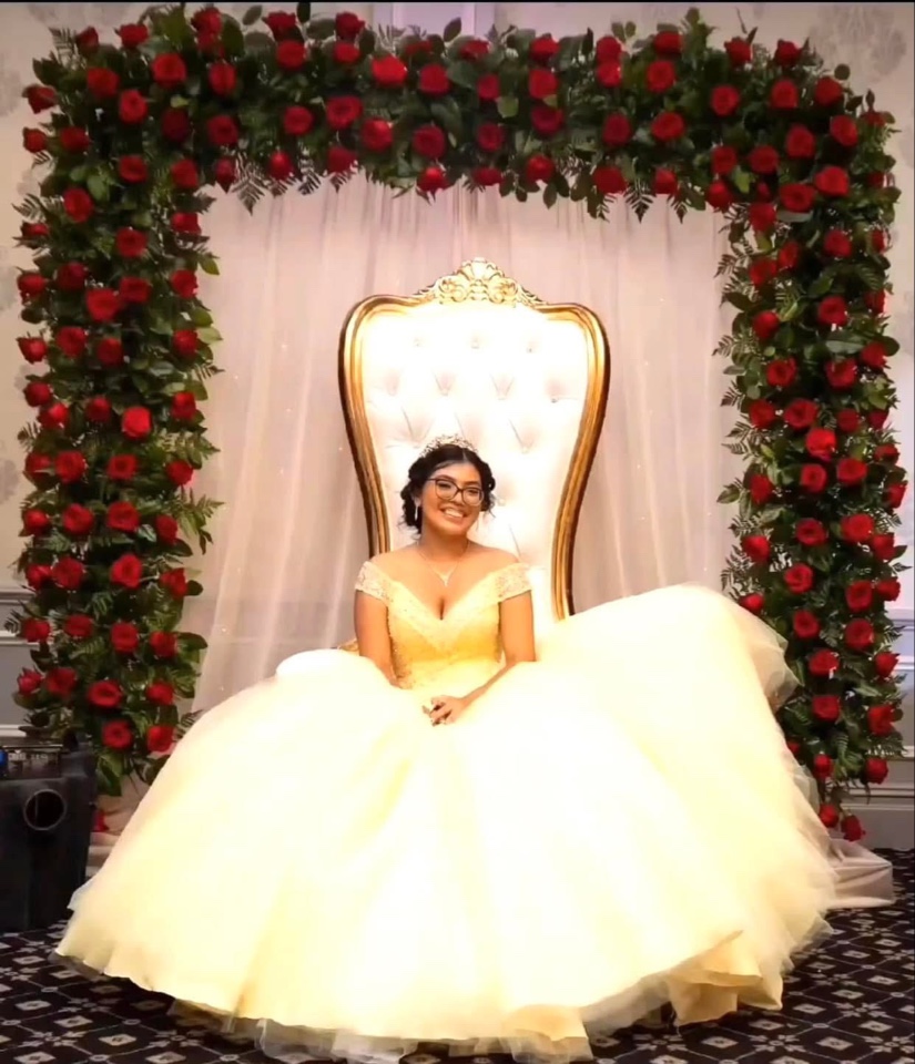 Quinceañera in a yellow gown on a throne chair beneath a grand red rose and greenery arch