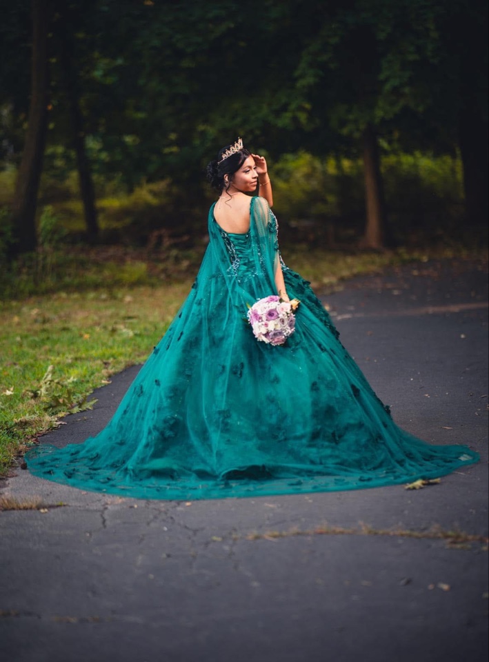 Quinceañera in an emerald ball gown and tiara carrying a lavender and blush bouquet down a garden path